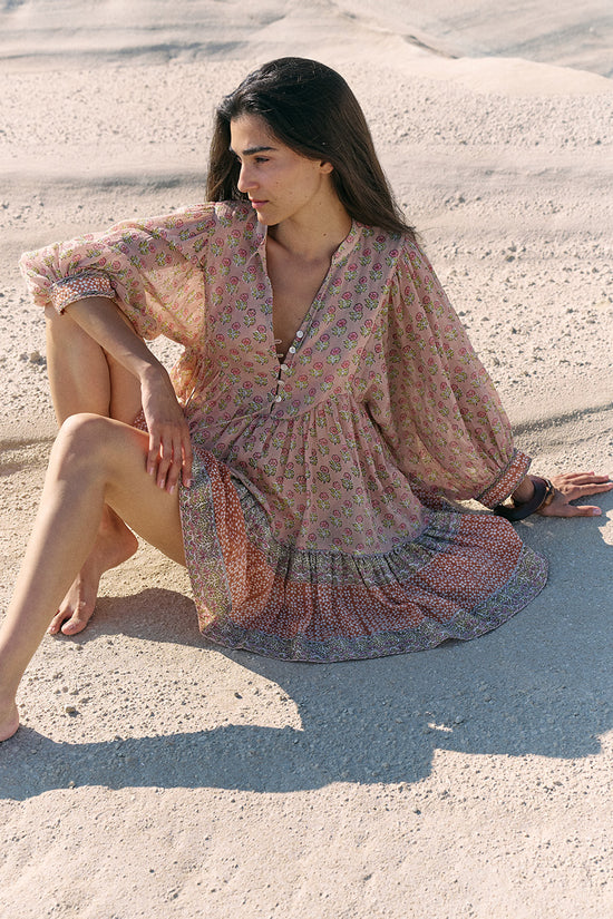 Woman seated on sand wearing the Daughters of India Bhoomi Mini Dress in Sunset, the peach floral print mini dress in a relaxed seated pose